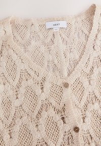 Kreemjas heegeldatud kardigan lillemustriga, millel on V-kaelus ja ees kaks pruunikat nööpi. Õhuke, tekstuurne kangas.