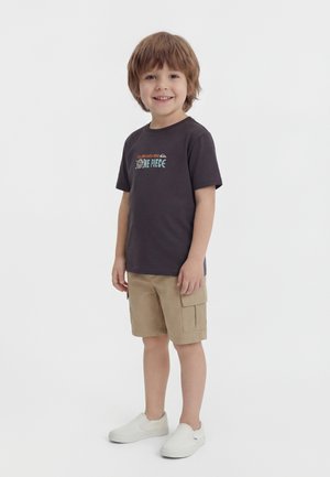 Smiling boy with light brown hair wears black One Piece graphic t-shirt, beige cargo shorts, and white slip-on shoes, standing on white background.