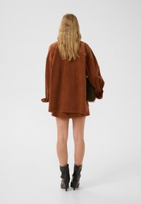 Chemise en velours côtelé marron oversize à manches longues, associée à une jupe assortie et des bottes noires pointues jusqu'aux genoux. Tissu lisse et texturé.