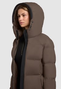 Frau in einer braunen Steppjacke mit großer Kapuze, teilweise ungezogen, um ein schwarzes Top mit sichtbarem Reißverschluss zu zeigen.