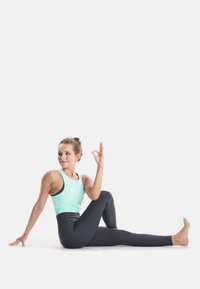 Svijetloplava atletska majica bez rukava i crne tajice visokog struka. Figura sjedi u yoga pozi, s podignutom rukom u gesti svjesnosti.