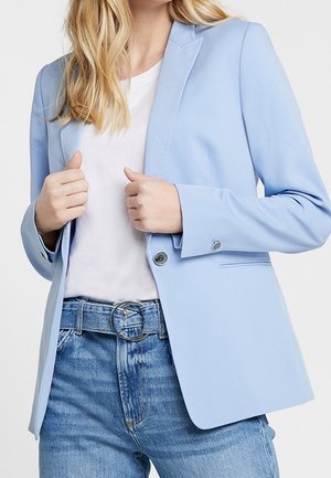 Mujer con blazer azul claro sobre camisa blanca, jeans azules y cinturón con hebilla redonda plateada, ajustando el frente del blazer con ambas manos.