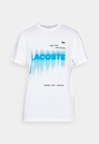 Vit bomull t-shirt med blå gradienttext som säger "LACOSTE" och "SE YOU ON COURT." Innehåller en grön krokodil-logotyp och svarta detaljer.