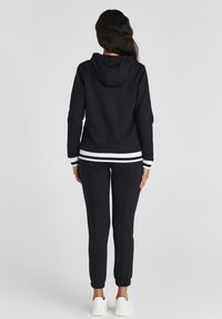 Černá mikina s nastavitelnou kapucí, bílé pruhované akcenty na manžetách a lemu; kombinováno s černými tepláky a bílými teniskami.