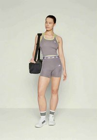 Žena v šedém a neonově zeleném sportovním oblečení, která drží černou tašku přes rameno, má na sobě bílé tenisky a pruhované ponožky, stojící na jednobarevném pozadí.