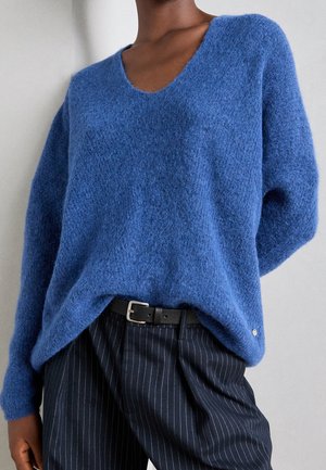 Persoon draagt een zachtblauwe v-hals trui, ingestopt in zwarte pantalons met krijtstrepen en een zwarte riem, tegen een neutrale achtergrond.