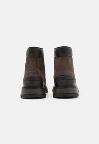 Stivali alla caviglia in suede marrone scuro con dettagli in pelle nera, suola in gomma piatta e cuciture rinforzate lungo le cuciture.