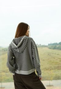 Szara i czarna bluza z kapturem w paski o krótkim kroju. Dziergane ściągacze na mankietach i brzegu, miękka faktura. Noszona z ciemnymi spodniami. W tle widok na zewnątrz.