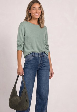 Femme portant un haut vert à manches longues et un jean bleu, tenant un sac à main vert foncé avec une expression souriante.