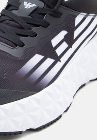 Fekete atlétikai cipő texturált anyaggal, fehér részletekkel és geometriai mintájú talppal, kontrasztos mintázatú oldalelemekkel.