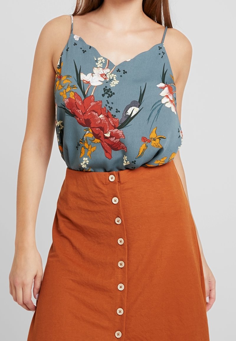 Blouse bleue à motif floral avec un décolleté festonné, associée à une jupe orange à boutons avec des boutons en bois et une texture lisse.