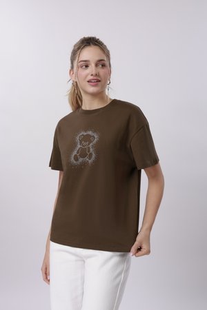 Jeune femme aux cheveux clairs attachés en queue de cheval, portant un t-shirt marron avec un motif ours en peluche scintillant, associé à un pantalon blanc sur un fond uni.