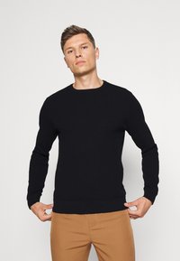 Suéter negro de punto con cuello redondo y mangas largas, con un patrón texturizado y puños y dobladillo acanalados. Llevado sobre pantalones color tostado.