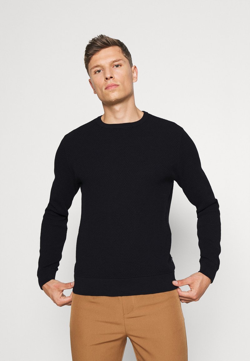 Suéter negro de punto con cuello redondo y mangas largas, con un patrón texturizado y puños y dobladillo acanalados. Llevado sobre pantalones color tostado.