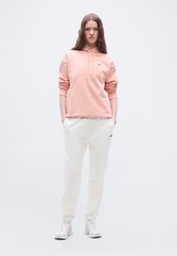 Ženska stoji z rokami v žepih, oblečena v svetlo roza kapuco, bele trenirke in bele superge, na enostavni svetli podlagi.