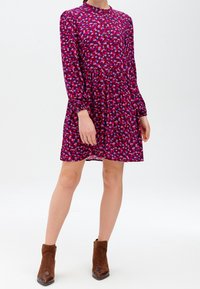 Długa, fioletowa sukienka z długim rękawem, zdobiona printem serc w odcieniach różu i niebieskiego, z zebranym w talii detalem i zapięciem na guziki. Noszona z brązowymi botkami za kostkę.