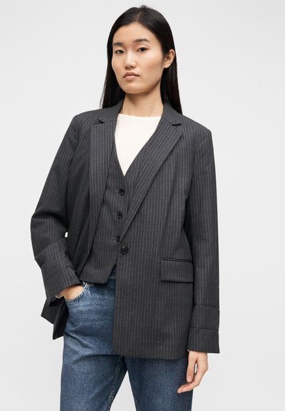 Blazer gris à fines rayures avec une coupe structurée, des revers à encoches et des poches avant, porté sur un gilet gris et un haut clair.