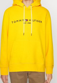 Sudadera amarilla con capucha y cordones blancos, que presenta el texto de logo "TOMMY HILFIGER EST. 1985" en negro. Material de algodón, con bolsillo tipo canguro.
