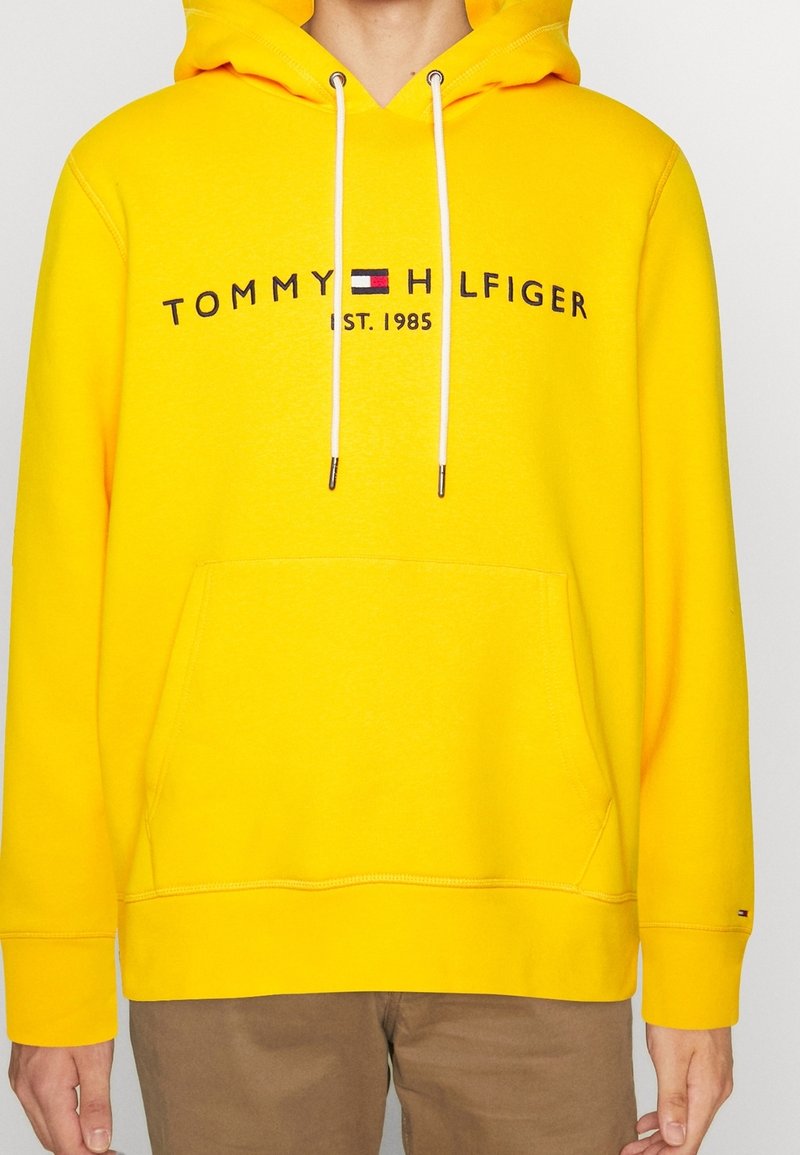 Sudadera amarilla con capucha y cordones blancos, que presenta el texto de logo "TOMMY HILFIGER EST. 1985" en negro. Material de algodón, con bolsillo tipo canguro.