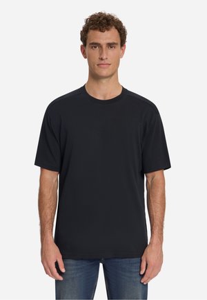 Man met krullend haar die een effen zwart t-shirt met korte mouwen en een blauwe spijkerbroek draagt, staand tegen een lichtgrijze achtergrond.