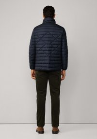 Mężczyzna stojący przodem do jasnoszarej ściany, ubrany w granatową pikowaną kurtkę, ciemnozielone spodnie i brązowe buty.