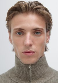 Mladý člověk se světle hnědými vlasy, modrýma očima a neutrálním výrazem, oblečený v šedém svetru na zip s vysokým límcem, na jednobarevném pozadí.