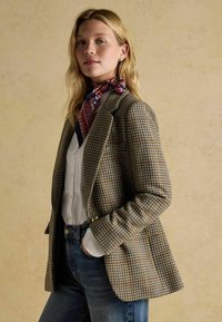 Hnedý houndstooth blazer so zlatými gombíkmi, nosený cez bielu košeľu a modré džínsy. Doplnkový červený a modrý vzorovaný šál.