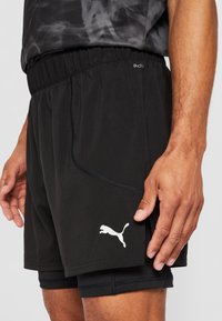 Mies, joka käyttää mustia Puma-urheiluhousuja, joissa on sisäinen puristuskerros, sekä kuviollista mustaa urheilusorttia, käsi levännyt sivulla.