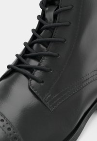 Melna ādas zābaks ar gludu apdari, noapaļotu pirkstu, tradicionālu auklu pievilkšanu un dekoratīvu šuvumu gar malām.