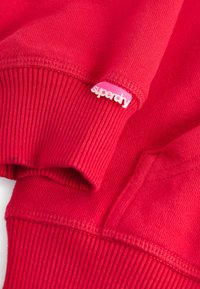 Červená mikina s ribovanými manžetami a lemem, se malou růžovou nášivkou loga Superdry na rukávu. Měkká, texturovaná látka.