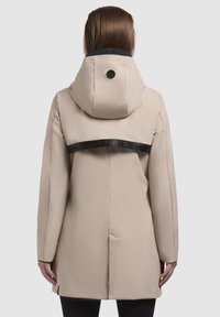 Veste imperméable beige avec capuche, sangle noire avec logo, texture lisse et coupe droite. Présente un logo sur la capuche.