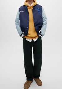 Person klädd i en marinblå varsityjacka med texten "Yale University", senapsgul huvtröja, vit skjorta, svarta byxor och bruna mockaskor.