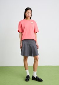 Roze katoenen T-shirt met korte mouwen en bloemenlogo, gecombineerd met een grijze geplooide rok. Het model draagt zwarte instappers en witte sokken.