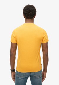 Gul kortærmet T-shirt med rund hals, lavet af blødt stof. Har en lige kant og design på bagsiden, sammen med blå jeans.