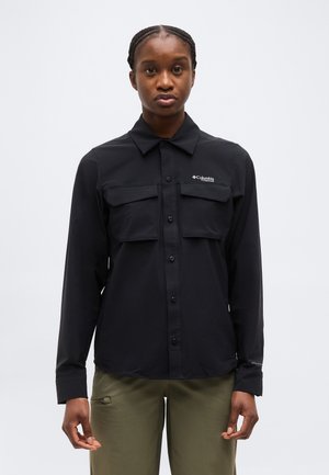 Άτομο που φοράει μαύρο πουκάμισο με κουμπιά και δύο τσέπες στο στήθος και χακί παντελόνι, στέκεται μπροστά σε ένα απλό φωτεινό φόντο.