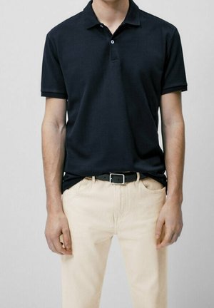 Polo shirt bleu marine avec patte de boutonnage à trois boutons, manches courtes et tissu uni. Porté avec un pantalon de couleur claire et une ceinture noire.