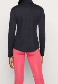 Czarna bluzka z długim rękawem i wysokim kołnierzem, dopasowany krój i gładka faktura, połączona z różowymi, przylegającymi legginsami. Brak widocznych elementów metalowych ani wzorów.