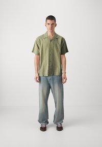 Camicia verde a maniche corte con un motivo sottile, abbinata a jeans larghi in denim chiaro e sneakers scure con lacci bianchi.