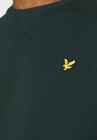 Suéter verde oscuro con textura acanalada, que presenta un pequeño logotipo de pájaro amarillo bordado en el lado izquierdo del pecho. Diseño de cuello redondo.