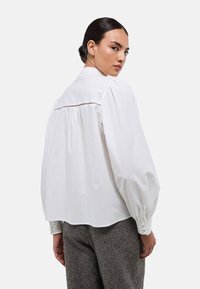 Blouse en coton blanc avec manches bouffantes, col rond et dos plissé. Présente des coutures délicates le long de l'encolure pour le détail.