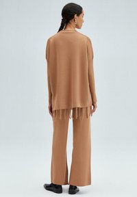 Tan oversized trui met franjes aan de onderkant, gecombineerd met breedvallende tan broek en zwarte schoenen. De stof lijkt zacht met een textuurafwerking.