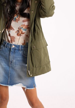 Femme portant une veste vert olive sur une chemise imprimée de chérubins, associée à une mini-jupe en denim à ourlet effiloché.