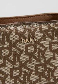 Surface de sac marron et beige à motifs avec des lettres répétées "DKNY" et un logo métallique doré "DKNY" centré sur un tissu texturé.
