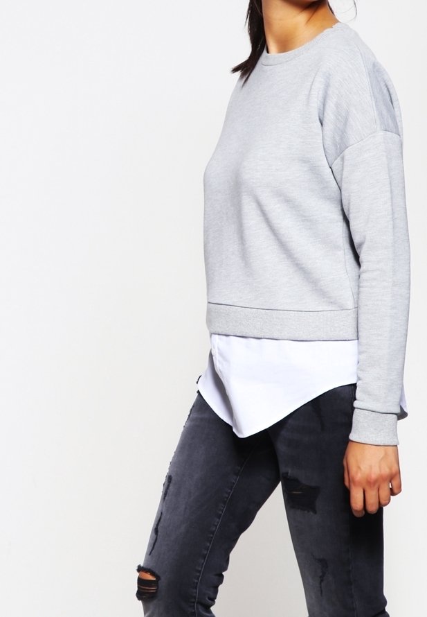 Mujer vestida con un sudadera gris claro superpuesta a una camisa blanca, con jeans ajustados rasgados negros, de pie contra un fondo blanco liso.