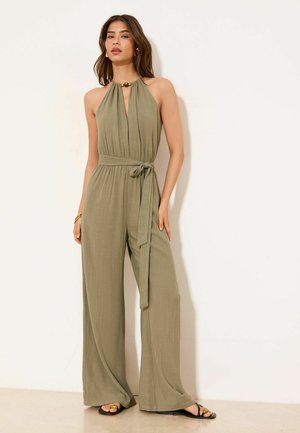 Woman wearing olive green halter-neck wide-leg jumpsuit with waist tie and black sandals against white background.