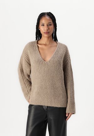 Beige gerippter Strickpullover mit tiefem V-Ausschnitt und langen Ärmeln, aus weichem, strukturiertem Stoff und entspanntem Schnitt, kombiniert mit schwarzen Lederhosen.