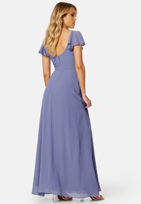 Maxi-jurk in zachte lavendelstof met fluttermouwen, een lage rugontwerp en een vloeiende silhouette. Heldergelakte sandalen maken de look compleet.