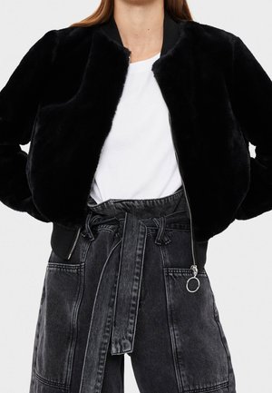 Femme portant une veste en fausse fourrure noire sur un t-shirt blanc et un pantalon en jean noir taille haute avec une ceinture nouée.