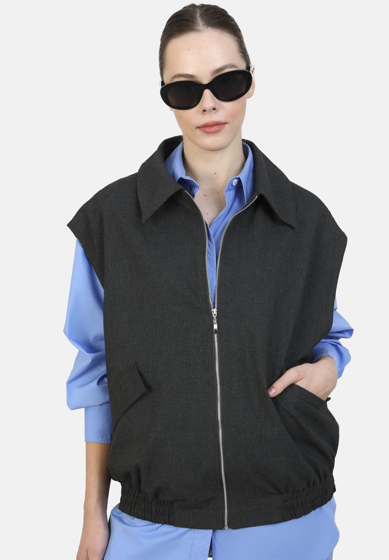 Giubbotto senza maniche grigio scuro con colletto, zip frontale, orlo elasticizzato e due tasche laterali, indossato sopra una camicia azzurra chiara.