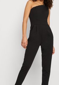 Combinaison noire asymétrique avec un corsage ajusté et une bordure texturée. Caractéristiques des jambes fuselées et des plis à la taille pour un look structuré.